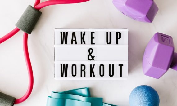 Top view closeup composition of wake up and workout inscription on light box surface among expanders and dumbbells and massage ball on white background
